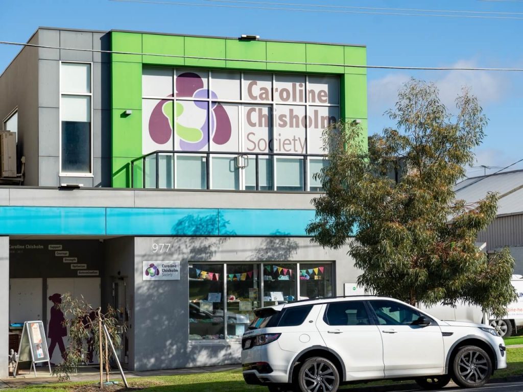 A modern two-storey building with green exterior on the second-floor displaying the Caroline Chisholm Society logo, and a ground floor with grey exterior featuring a wide window displaying essential baby clothes and goods.